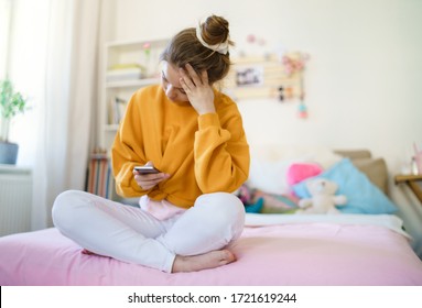 Sad Young Female Student Sitting On Bed, Using Smartphone.