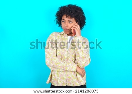 Sad young girl with afro hairstyle wearing floral shirt over blue background talking on smartphone. Communication concept.