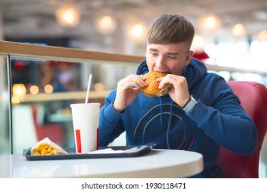 Sad Young Guy Eats On A Food Court, Fast Food. The Man Eats A Bites Burger And Drinks Soda Water. Bad Food, Street Food.