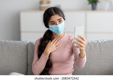 Sad Young Indian Lady In Protective Mask On Sofa Makes Selfie On Smartphone Or Video Call In Living Room Interior. Social Distance And Communication Through Modern Device During Covid-19 Quarantine