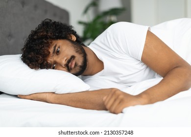 Sad young indian man in pajamas laying in bed, thinking about something, feeling down, reluctant to wake up in the morning, closeup shot. Depression, sadness, frustration for millennials
