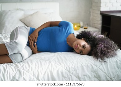 Sad Young Latina Woman With Menstrual Pain. Black Girl Having Cramps During Period And Lying On Bed At Home