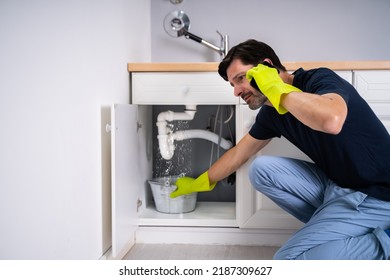 Sad Young Man Calling Plumber In Front Of Water Leaking From Sink Pipe
