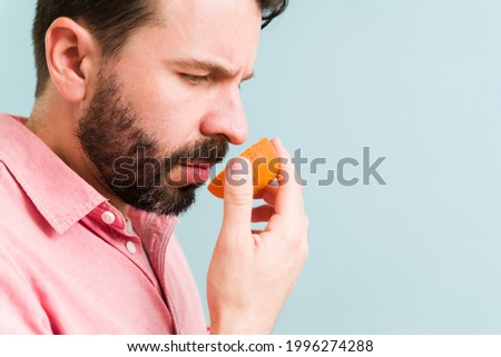 Sad young man holding an orange fruit in front of his nose and trying to smell it. Latin man with anosmia illness