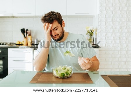 A sad young man looks longingly at a salad, holds a fork with greens in his hand, and a plate of salad in front of him. An exhausting diet haunts, hates healthy food. Stop diet.