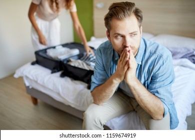 Sad young man sitting on bed and thinking of what to do while his wife going to leave
