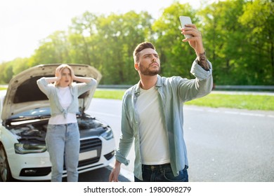 Sad young man standing on road next to his broken car together with terrified girlfriend, raising hand with mobile phone, looking for network signal, trying to call roadside assistance. No connection