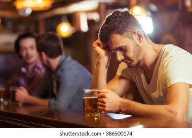 Sad young man in white t-shirt is looking at glass of beer while sitting at bar counter in pub, in the background two other men - Powered by Shutterstock