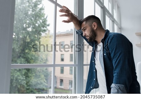 Sad young man with worried stressed face expression looking out window. Obsessive compulsive, adhd, anxiety disorders. Unhappy european handsome man stands, presses hand to glass in kitchen interior