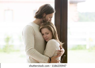 Sad young married couple embracing standing in living room opposite window at home. Sorrowful wife and husband feels unhappy, thinking about problems in relations, miscarriage, misbirth or infertility
