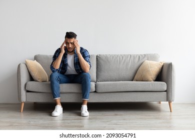 Sad young Middle Eastern man suffering from headache or migraine, touching head massaging temples. Stressed guy sitting on couch with painful face expression feeling terrible weakness or depression - Shutterstock ID 2093054791