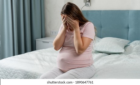 Sad young pregnant woman holding big pillow and sitting on bed. Concept of maternal and pregnancy depression