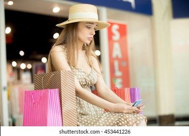 Sad Young Pretty Woman Sitting In Shopping Mall With Shopping Bag Around Thinking. She Is Holding Credit Cards Looking Upset As No Money Left. Buying Too Much On Expensive Shopping Time Concept