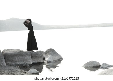 Sad Young Woman In Black Vintage Dress Stands By The Sea Being Tortured By Sorrow. 19th Century Novel. Copy Space. Black And White Shot.