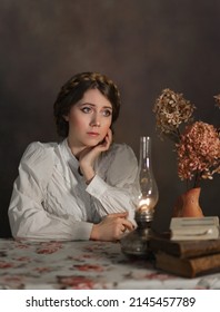 Sad Young Woman With Braids In Old Fashioned Vintage Blouse Sitting At The Table With Old Lamp, Books And Flowers