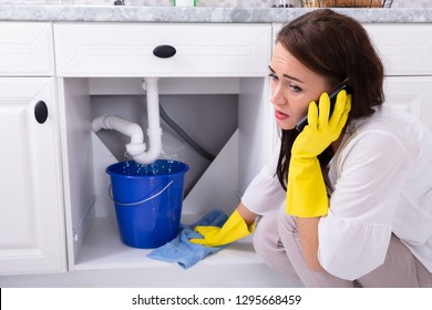 Sad Young Woman Calling Plumber In Front Of Water Leaking From Sink Pipe