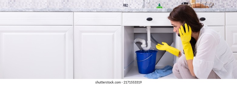 Sad Young Woman Calling Plumber In Front Of Water Leaking From Sink Pipe