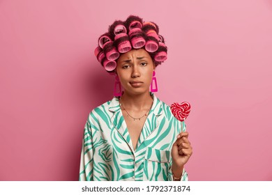 Sad Young Woman Gets Hair Permed, Looks Unhappily At Camera, Wears Hair Rollers, Dressed In Casual Clothes, Holds Sweet Lollipop, Spends Time On Herself, Caring About Her Beauty, Isolated On Pink Wall