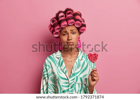 Sad young woman gets hair permed, looks unhappily at camera, wears hair rollers, dressed in casual clothes, holds sweet lollipop, spends time on herself, caring about her beauty, isolated on pink wall