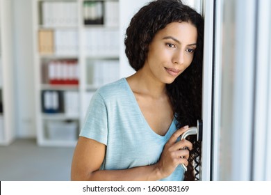 Sad Young Woman Looking Longingly Out Of A Glass Door At The Office With A Quiet Smile And Head Resting On The Door