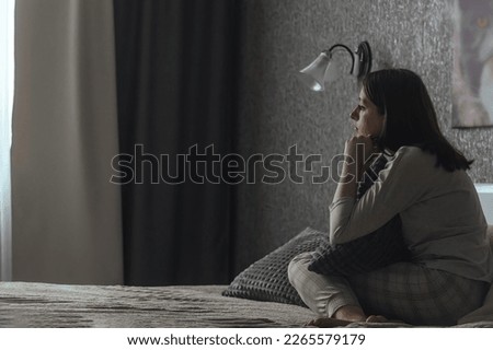 A sad young woman in pajamas with seasonal affective disorder or depression sits alone on the bed and looks out the window. The concept of winter depression due to lack of sunlight