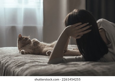 Una triste joven con trastorno afectivo estacional yace sola en la cama y mira por la ventana, al lado de un gato doméstico. Concepto de depresión de invierno debido a la falta de luz solar, foco selectivo