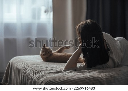 A sad young woman with seasonal affective disorder lies alone on the bed and looks out the window, next to a domestic cat. Concept of winter depression due to lack of sunlight, selective focus
