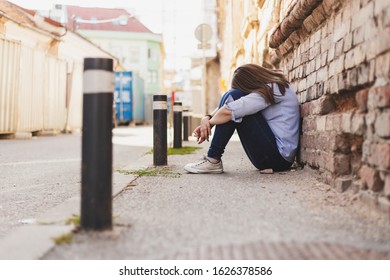 Sad Young Woman Sitting Alone On The Concrete In The City With Her Head On Her Knees - Stressed Casually Dressed Female Worker Feeling Anxiety Outside - Concept Image For Domestic Abuse