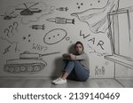 Sad young woman sitting near wall with drawn tank, helicopters and missiles. Horrors of war