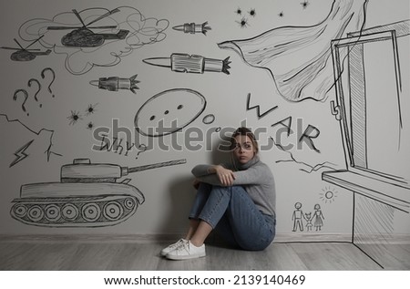 Sad young woman sitting near wall with drawn tank, helicopters and missiles. Horrors of war