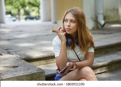 Sad young woman is sitting on the stairs of garden pavilion with ice cream and smartphone in hands. Pensive beautiful blonde female about 20 years old has a rest in public park.