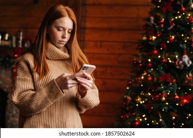 Sad Young Woman Typing Message On Mobile Phone While Self-isolating During The Winter Holidays At Room With Festive Interior. Concept Of Celebrating Christmas And New Year At Alone While Lockdown.