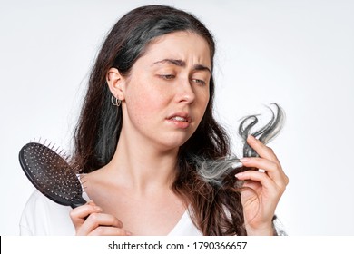 A Saddened Brunette Woman Holds A Comb With Her Hair Falling Out And Looks At The Gray Tips Of Her Hair. White Background. The Concept Of Hair Loss And Gray Hair