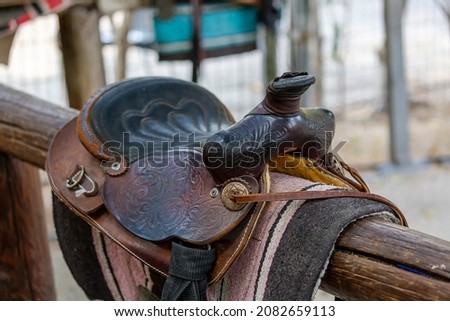Saddle brown on fence in shallow depth of field. Sport leather saddle, harness for horses on daylight. Western horse saddles on a rack,  ready for dressage training. Equestrian sport background. 