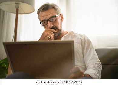 Sadly Old Man Sitting Alone On Sofa Looking At Picture Frame And Thinking Of Someone In The Picture. Concept Elderly Missing Someone That Pass Away After Covid-19 Pandemic.