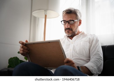 Sadly Old Man Sitting Alone On Sofa Looking At Picture Frame And Thinking Of Someone In The Picture. Concept Elderly Missing Someone That Pass Away After Covid-19 Pandemic.