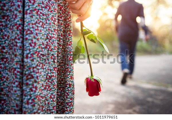 Sadness Love in Ending of Relationship\
Concept, Broken Heart Woman Standing with a Red Rose on Hand,\
Blurred Man in Back Side Walking away as background\
