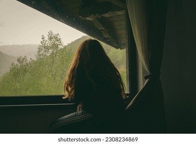  Sadness, melancholy, nostalgia - concept. A woman looks out the window on a rainy day at the mountain cabin.