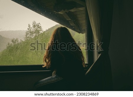  Sadness, melancholy, nostalgia - concept. A woman looks out the window on a rainy day at the mountain cabin.