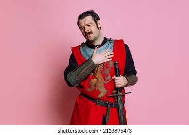 Sadness. One Young Man, Medieval Warrior Or Knight Wearing Wearing Armor Clothing Posing Isolated Over Pink Background. Model With Wounded Face. Comparison Of Eras, History, Funny Meme Emotions