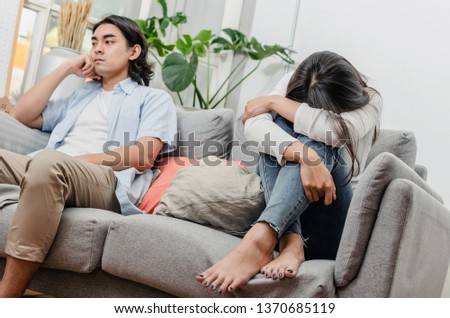 sadness young asian wife having quarrel and sitting on sofa after fight with husband behind her in house interior together, upset couple, love, divorce couple, family issues and relationship concept