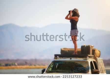 Safari activities in Zambia: Silhouette of a Fit Woman standing on the roof of safari car, observing Zambezi River Nature through binoculars. Lower Zambezi National Park, Zambia, Africa.