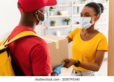 Safe Delivery. Black Lady Taking Delivered Parcel Box From Male Courier Wearing Face Mask And Gloves Standing At Home. Deliveryman Shipping Package To Customer Woman. Selective Focus