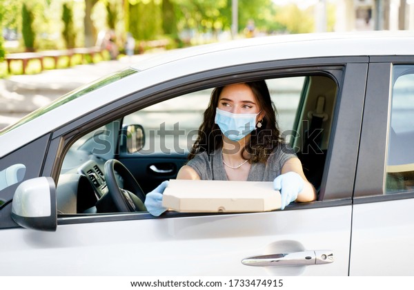 Safe food delivery from pizzeria to car during\
quarantine covid-19. Attractive young girl in blue medical mask and\
gloves gets pizza in cardboard box. Delivering food by car\
according social distance.