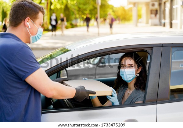 Safe food delivery from pizzeria to car during\
quarantine coronavirus. Attractive business woman in medical mask\
and gloves gets pizza in cardboard box. Delivering by car according\
social distance