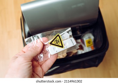 Safe Handling And Management Of Rapid Antigen COVID-19 Testing Waste Concept. Person Hand Throw Away Pieces Of Antigen Test Kit Sealed In Plastic Bag, Biohazard Symbol. Home Use.