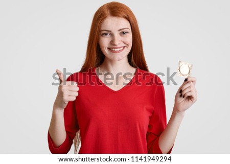 Safe pleaure and protection concept. Glad redhaired female with freckled skin holds condom, shows ok sign, dressed in red clothes, isolated over white background, preserves herself from pregnancy