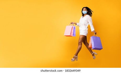 Safe Shopping. Black Woman In Medical Face Mask Running In Mid-Air Carrying Colorful Shopper Bags Posing Over Yellow Background. Studio Shot, Panorama With Copy Space For Text - Shutterstock ID 1832649349