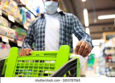 Safe Supermarket Shopping. African Man Disinfecting Shopping Cart Trolley Using Sanitizer And Paper Napkin For Virus Protection Buying Food In Grocery Store, Wearing Mask. Cropped, Selective Focus