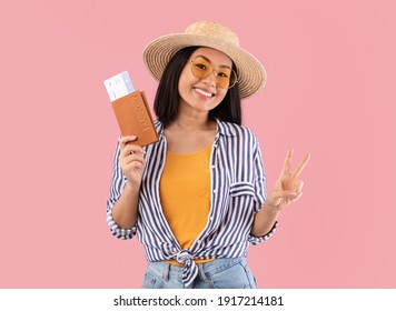 Safe Travels. Portrait of cheerful asian lady holding passport and tickets, gesturing peace v sign, positive tourist woman posing in stylish straw hat and sunglasses over pink studio background